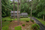 Two-story house located at 70 Windsor Court, Henderson NC, with a gray roof and yellow siding, surrounded by tall pine trees and greenery, featuring a winding driveway.