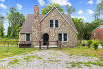 A quaint stone cottage with a steeply pitched roof and an arched front door, located at 1203 Dorsey Avenue, Henderson NC, is set against a backdrop of greenery under a partly