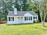 A single-story white house with black shutters and a front porch, featuring a chimney and surrounded by a lawn with trees, located at 1602 Bane Avenue, Henderson NC.