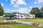 A single-story white house with a metal carport sheltering two vehicles, located at 151 Southerland Mill Road, Henderson NC, is set against a backdrop of green lawn and trees under a