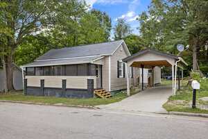 A single-story house at 920 David Street, Henderson NC, with beige siding and a dark trim, featuring a carport on the right and surrounded by trees, with a front yard mailbox by the