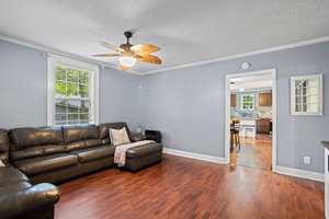 A clean and well-lit living room with a dark brown leather sofa, hardwood floors, and light gray walls, featuring a ceiling fan above and a view into an adjoining room with a kitchen counter.