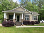 A single-story suburban home in Henderson NC, with beige siding, a dark roof, and white trim featuring a front porch with two columns, landscaped with burgundy shrubs, and surrounded by a