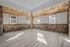 An empty room with stone and wood wall accents, grey flooring, and multiple windows allowing natural light.