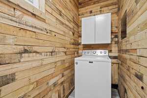 A compact laundry room with rustic wooden plank walls featuring a white top-loading washing machine and a white dryer stacked under a cabinet.