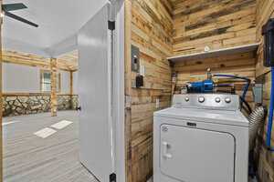 A laundry room corner with a white top-loading washing machine and a blue utility pump installed above it, featuring rustic wooden wall paneling and a view into an adjoining room with a stone fireplace.