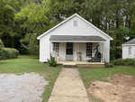 A small white house at 556 High Street, Henderson NC, with a gabled roof featuring a front porch with an exercise bike and a couple of chairs, surrounded by greenery and a partially paved