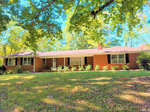 A single-story brick house at 1204 David Avenue, Henderson NC, with a sloping front yard and mature trees casting shadows on the lawn.