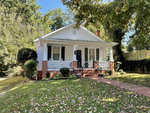 A quaint, white single-story house at 226 W Belle Street, Henderson NC, with a red brick pathway, flanked by trees and fallen leaves, on a sunny day.