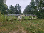 An abandoned single-story house at 295 Marigold Lane, Henderson NC, with a yellow exterior, overgrown with vegetation, set against a backdrop of dense trees.