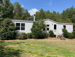 A single-story white manufactured home with a chimney and a small set of wooden stairs, situated among trees and vegetation under a partly cloudy sky at 71 Plum Lane, Henderson NC.