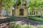 A stone pathway leads to a rustic wooden door set within a stone archway entrance of the house at 550 Deepwood Drive, Henderson NC, surrounded by lush green trees and foliage.