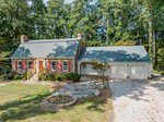 A single-story red brick house located at 156 Cheatham Mabry Road, Henderson NC, with white garage doors, a chimney, and a landscaped front yard featuring a stone pathway and circular