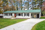 A single-story residential home at 1508 Lynne Avenue, Henderson NC, with a green metal roof, white siding, and a red front door, set against a backdrop of autumn trees.