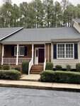 A single-story brick-front house with a small porch and white door, located at 2060 Fernwood Drive Unit 2, Henderson NC, surrounded by shrubbery, with a backdrop of tall