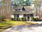A two-story house at 306 S B Harrison Lane, Henderson NC, with beige siding and white trim, surrounded by a wooded area with a clear sky overhead.