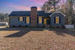 A single-story, charcoal-gray house with white trim and brick accents at 79 Holly Circle, Henderson NC, featuring a large chimney and a fenced yard, cast under the shadows of bare trees.