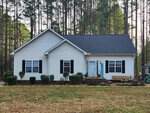 A single-story white house at 91 Meridian Way, Henderson NC, with a blue door, flanked by symmetrical windows and surrounded by tall pine trees.