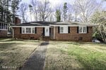 A single-story brick house at 205 Edgewood Drive, Henderson NC, with a black shingled roof, featuring a white front door and surrounded by a yard with trees.