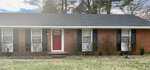 A single-story brick house with a red door and a shingled roof, located at 224 White Oak Drive, Henderson NC, is fronted by a lawn with scattered grass and bare shrubs