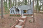 A single-story gray house at 107 Orchard Road, Henderson NC, with white trim and a red door, surrounded by tall pine trees. The house is accessed by a brick staircase leading up to a