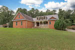 A brick single-story house with a gabled roof and dormer windows, featuring a large front lawn and a clear blue sky. a covered porch with pillars and a driveway to the left are also visible.