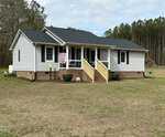 A one-story white house with a gabled roof, front porch with wooden steps, and several windows, set on a large lawn bordered by a forest at 242 Edwards Road, Henderson NC. A