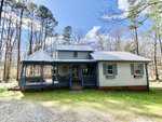 A quaint blue house with a gray roof, located at 358 Pool Rock Shores Lane in Henderson, NC, featuring a wrap-around porch and a screened gazebo, nestled in a wooded area with