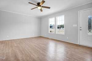 Empty living room with gray walls and light wooden flooring. the room features a ceiling fan, a front door with decorative glass, and two large windows overlooking a street.