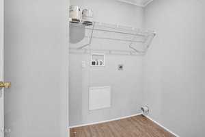 A small, empty laundry room with white walls, a wooden floor, and a high-mounted wire shelf. there is a closed white door, a water heater, and utility panels on one wall.