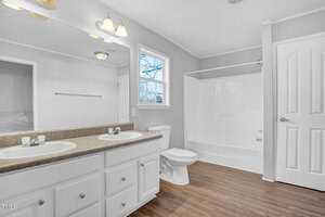 A bright bathroom featuring white cabinetry with dual sinks, a large mirror, a bathtub-shower combo, a toilet, and a window providing natural light. the flooring is wooden, and the walls are painted white.