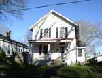 A two-story white house at 128 Carolina Avenue, Henderson NC, with a covered front porch, black shutters on the windows, and a small front yard. The house has a visible chimney and