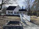 A small white house at 205 W Andrews Avenue, Henderson NC, with a sloped black roof and a porch, featuring a long white fence leading to the front door. The yard includes a bare