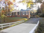 A brick single-story house at 227 Willowood Drive, Henderson, NC with white trim, surrounded by trees with autumn foliage. A white fence runs along the driveway leading to the house.