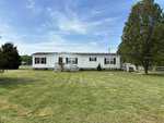 A white mobile home with a gray roof, featuring a front porch and stairs, positioned at 8340 Nc-39, Henderson NC on a large grassy lot with scattered trees and