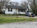A small, white, single-story house at 1402 S Williams Street, Henderson NC with a front porch and a black car parked in the driveway. There's a grassy area and bare trees
