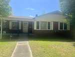 A one-story brick house with white siding and a covered entrance, located at 804 Eastside Drive, Henderson NC, featuring a small lawn and trees in the background on a sunny day. There is
