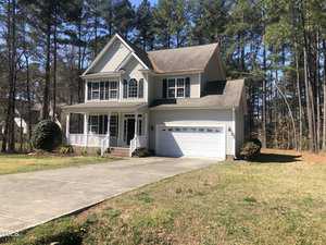 A two-story house with a white exterior and blue shutters, featuring a front porch and an attached two-car garage, surrounded by tall pine trees under a clear sky.