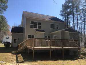 A two-story beige house with a large wooden deck, surrounded by tall pine trees on a clear sunny day. the house has black shingles and several windows, located in a suburban area.