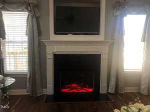 A cozy living room with an electric fireplace glowing red beneath a mounted television, flanked by two large windows draped with elegant gray curtains.