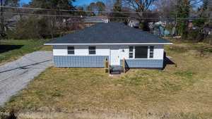 Aerial view of a small, white single-story house with a dark shingled roof, surrounded by a lawn with a gravel driveway, under clear blue skies.