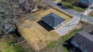 Aerial view of a small grey house with a white trim and a gable roof, surrounded by a large dry grass yard and located at a street intersection, with neighboring homes on either side.