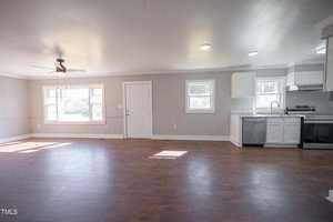 Spacious, well-lit room featuring dark hardwood floors, white walls, and a modern kitchen with white cabinets and granite countertops. there's a ceiling fan above and multiple windows providing natural light.