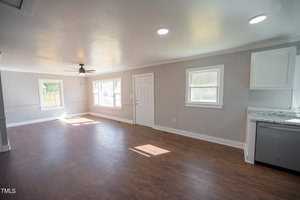An empty, sunlit room with hardwood floors, white and gray walls, a ceiling fan, a door, two windows, and a kitchen counter with cabinets on the right.