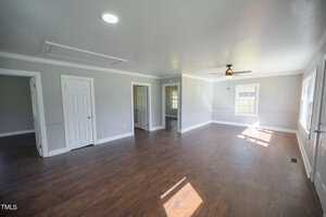 A spacious, empty room with polished wooden floors, white walls, and multiple doors. features include a ceiling fan, overhead lights, and a window letting in natural light.