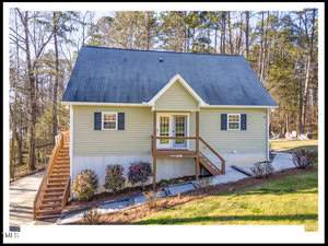 Aerial view of a two-story house with beige siding and a dark grey roof. the house features a front porch, stair access from the side, and is surrounded by trees and a landscaped yard.