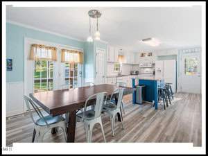 A bright, spacious kitchen and dining room with white walls, a dark wood dining table, metal chairs, a blue kitchen island with stools, and light wood flooring.