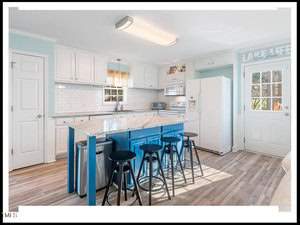 Bright, modern kitchen with white cabinetry, a granite countertop island with blue stools, subway tile backsplash, and hardwood floors. bright natural light floods in through the windows.