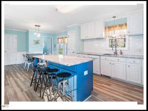 A bright, modern kitchen with white cabinetry and a vibrant blue island featuring bar seating. the room has light wooden flooring and pale blue walls, with natural light streaming in.