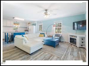 Bright and airy living room with white and blue decor, featuring a sectional sofa, a tv, and an open kitchen with bar stools in the background.